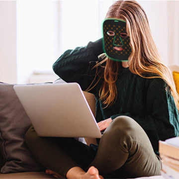 A woman using a laptop with a glowing mask on in a cozy indoor setting